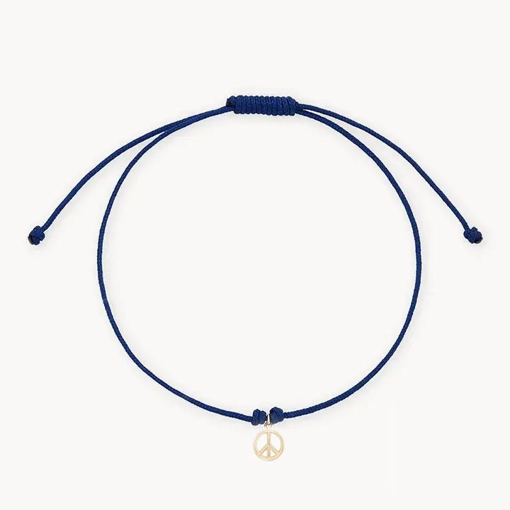 The tie dye moonchild contemplation cord bracelet by bluboho features a minimalist blue cord with an adjustable knot and a small 10k yellow gold peace sign charm hanging from the center. The bracelet’s ends are designed to dangle loosely against a plain white background.
