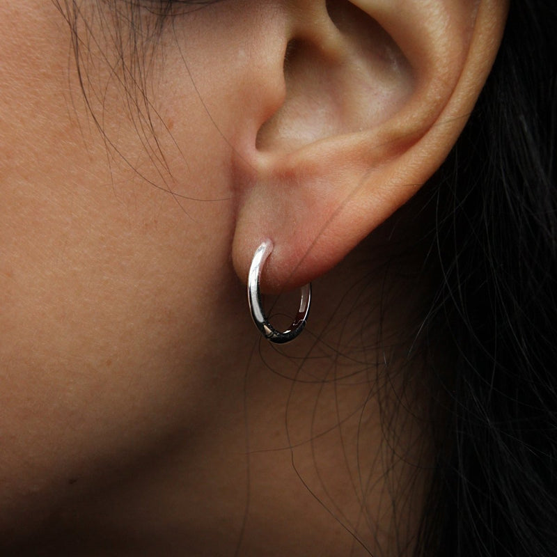 Close-up of a person’s ear adorned with the bluboho dagger medium hoop silver earring. Strands of dark hair frame the ear, while the background remains blurred, emphasizing the earring and ear.