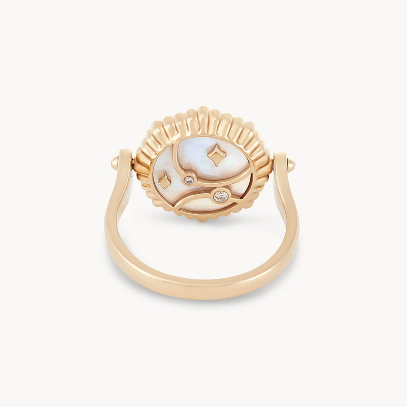 The moon phase flip enamel ring by bluboho is crafted in 14k yellow gold and features a unique circular face with mother of pearl and enamel details, accented with small diamonds set in an abstract design. The ring boasts a looped band and an intricate scalloped edge around the face, all set against a white background.