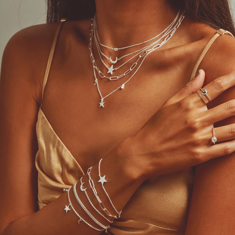 A person wearing a gold satin camisole adorned with multiple layered silver necklaces featuring stars, moons, and chains. Their left arm, crossed over their torso, displays matching sterling silver bluboho stella star bracelet and a ring. The background is plain and softly lit.