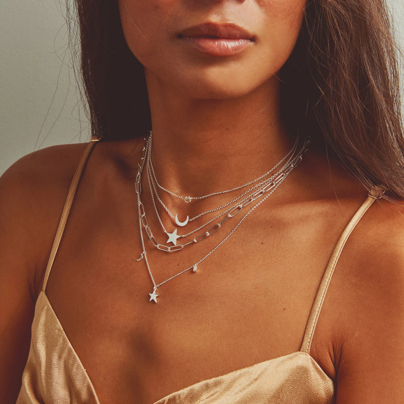 A close-up of a woman wearing multiple layered necklaces, including the "stella star necklace silver" by bluboho. Adorned with various charms, such as stars and a crescent moon, she is dressed in a silky beige spaghetti-strap top. The photo beautifully highlights both the intricate jewelry details and the texture of the fabric.