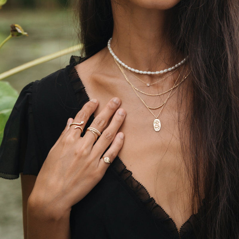 Close-up of a woman wearing multiple layered necklaces and rings. She has long, dark hair and is dressed in a black, sleeveless, V-neck top with lace trim. Her hand, adorned with the Serpent Revival Diamond Cuff Ring in 14k Yellow Gold by bluboho, rests on her chest, showcasing the jewelry against her skin.