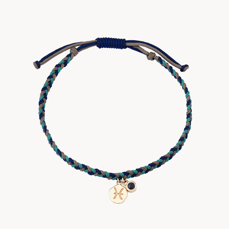 A bluboho pisces zodiac water element cord bracelet - 10k yellow gold, blue sapphire, cord featuring various shades of blue, teal, and gray, with an adjustable navy blue pull closure. The bracelet has two small charms: one circular gold charm with a Pisces zodiac symbol and another in the shape of an eye.