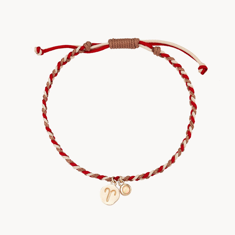 A bluboho aries zodiac fire element cord bracelet - 10k, opal, cord featuring red, tan, and white strands with an adjustable knot closure. It has two gold charms; a round one with an engraved zodiac sign and a smaller round one. The bracelet rests on a white background.