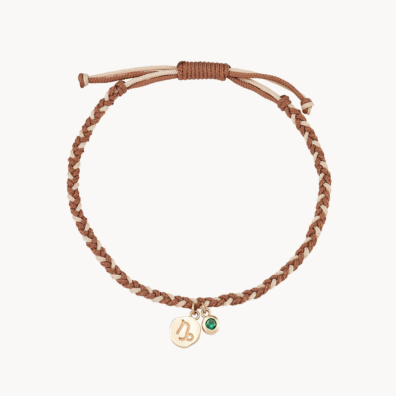 A brown and beige braided cord bracelet with an adjustable closure. It features two small hanging charms: a gold circle with a symbol etched on it, and a round, green gemstone by bluboho.