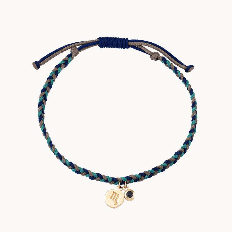 A scorpio zodiac water element cord bracelet - 10k yellow gold, blue sapphire, cord by bluboho with blue and turquoise threads and an adjustable closure. The bracelet features two small gold-colored charms: one circular with an engraved symbol and the other a tiny stone.