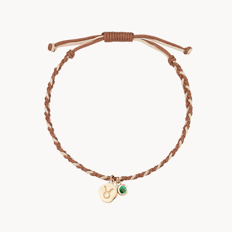 A brown woven cord bracelet with an adjustable sliding knot features two small gold charms: one engraved with a Taurus zodiac symbol and the other adorned with a green gem. This is the taurus zodiac earth element cord bracelet - 10k, emerald, cord by bluboho.