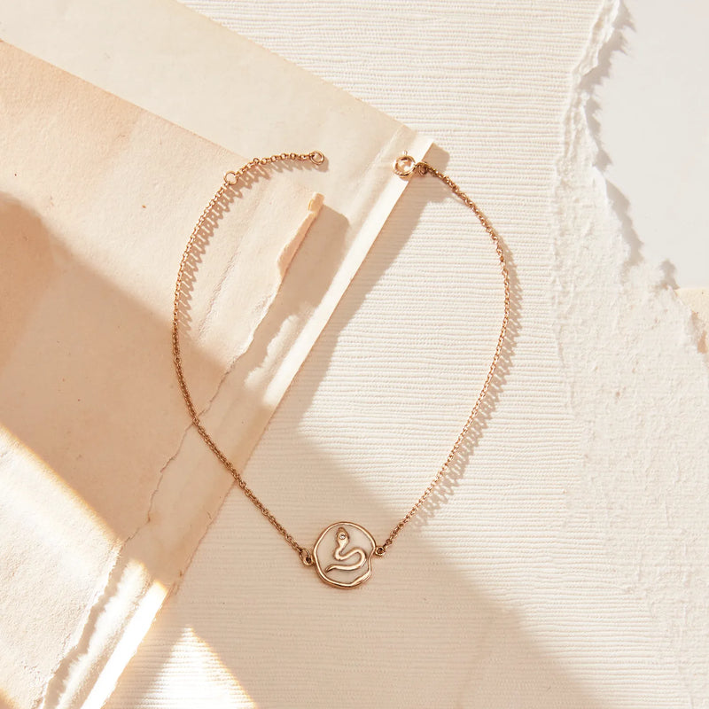 The revival snake enamel bracelet by bluboho, crafted in 14k yellow gold and accented with a diamond, is delicately placed on a textured white surface. Surrounding the bracelet are various torn pieces of beige and cream-colored paper, casting soft shadows in the natural light.