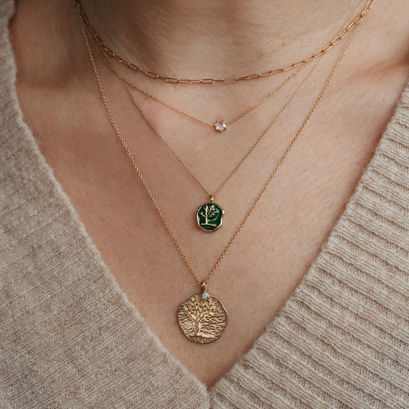 Close-up of a person's neck wearing three layered necklaces. The top necklace is a delicate gold chain featuring the serendipity October birthstone charm in 10k yellow gold with an opal by bluboho, the middle one has a green medallion with a tree design, and the bottom one features a larger gold medallion with another tree design.